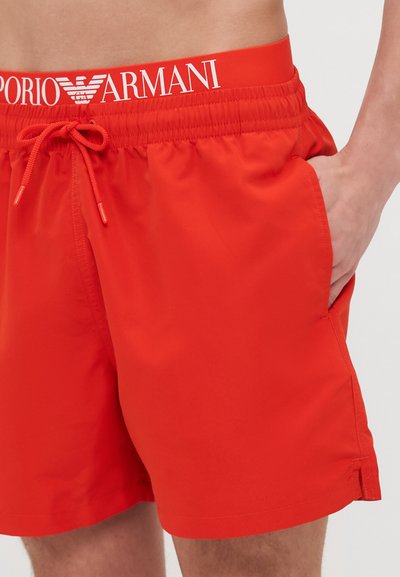 Červené plavky s elastickým pásom a šnúrkou, s bielym logom "Emporio Armani". Krátky strih so bočnými vreckami.