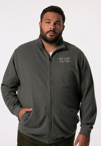 Chaqueta de forro polar con cremallera en verde oscuro, presenta un acabado texturizado, cuello elevado y la marca 'M 1997' en el pecho. Bolsillos en los laterales.