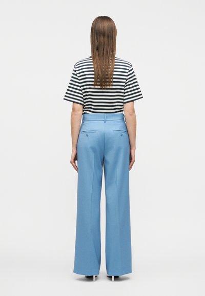 Άτομο που στέκεται με την πλάτη γυρισμένη φορώντας μια μαύρη και λευκή ριγέ μπλούζα, μπαλωμένη μέσα σε ανοιχτόχρωμο μπλε παντελόνι φαρδιάς γραμμής με κουμπωτές πίσω τσέπες.