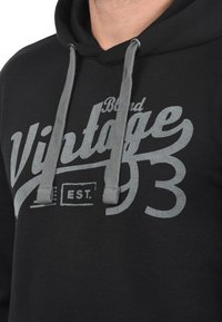 Sudadera negra con cordones grises. Presenta una gran impresión de estilo vintage en gris, con el texto "Brand Vintage 93" y "EST." en la parte delantera.