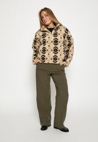 Béžový fleecový pulóver s čiernym a hnedým geometrickým vzorom; má zipsový golier, bočné vrecká, kombinovaný s olivovými nohavicami.