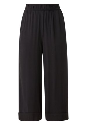 Žena oblečená v bílém pleteném topu bez rukávů, širokých černých kalhotách na zkrácenou délku a bílých teniskách, stojící s rukama v kapse.