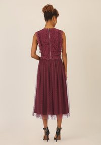 Bordeauxfarbenes Kleid mit Spitzenoberteil und gewellten Kanten, geschichteter Tüllrock mit Punkten, Reißverschlussverschluss hinten, getragen mit schwarzen Absätzen.