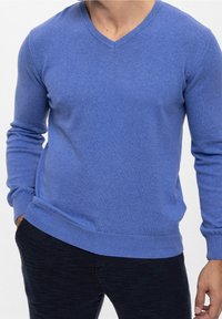 Homme portant un pull bleu à col en V à manches longues et un pantalon texturé foncé, une main dans la poche, sur un fond blanc uni.