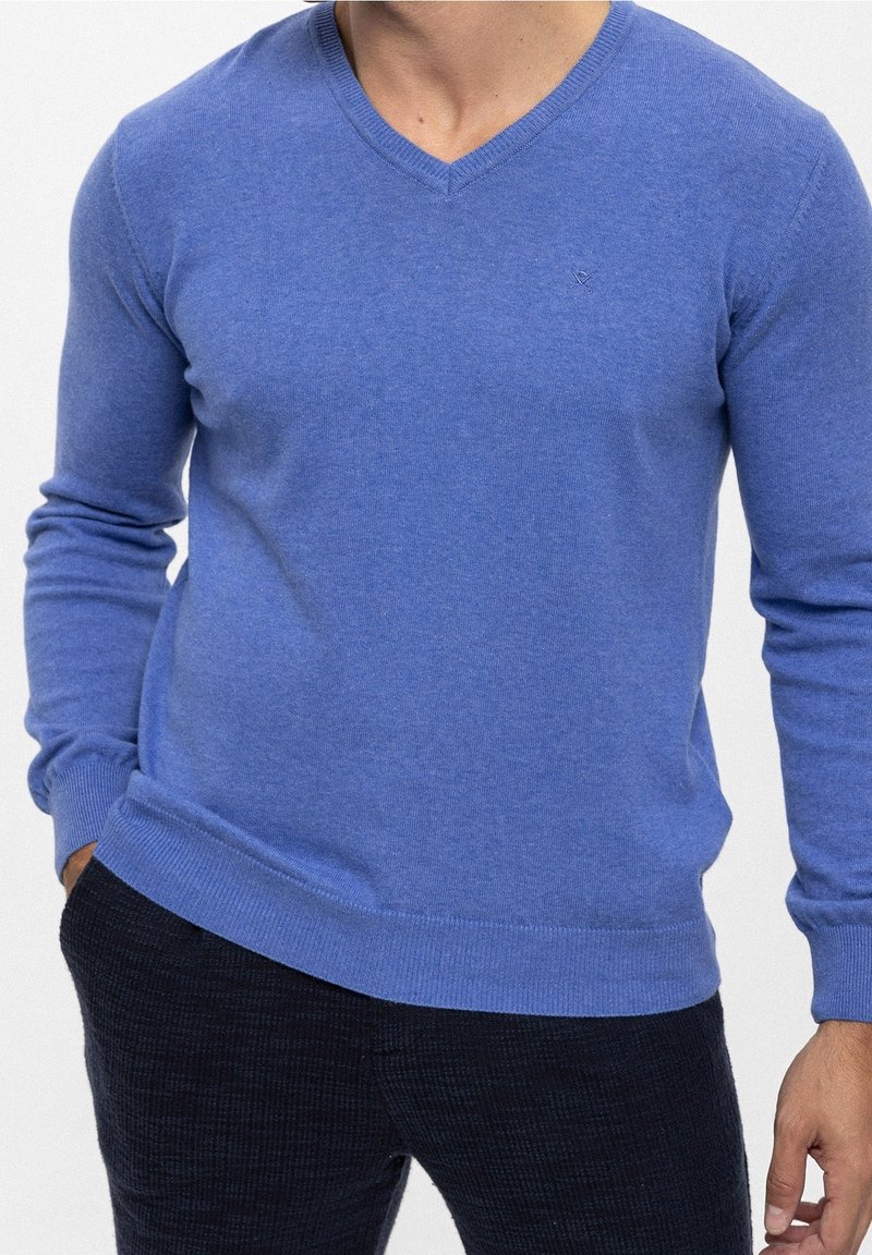 Homme portant un pull bleu à col en V à manches longues et un pantalon texturé foncé, une main dans la poche, sur un fond blanc uni.