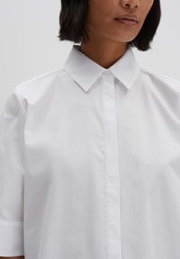Camicia bianca con colletto a punte e maniche corte, realizzata in tessuto liscio, con chiusura frontale a bottoni e un design classico e essenziale.