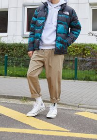 Giacca imbottita nera e verde acqua, abbinata a un felpa grigia, pantaloni beige morbidamente aderenti e sneakers alte bianche con accenti neri.