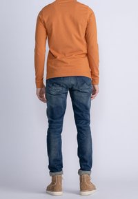 Långärmad orange bomullsskjorta, blå denimjeans, mockastövlar i tan. Synliga sömdetaljer och design på bakfickan på jeansen.