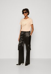 Perzik, cropped t-shirt met "GUESS" logo. Zwarte, glanzende, wijde cargo pants met zakken. Zwarte, puntige hoge hakken. Ronde zonnebril.