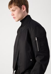 Black bomber jacket with ribbed collar, zippered arm pocket, slanted side pockets, and smooth matte texture. Silver zipper detail at front.