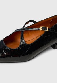 Chaussure en cuir verni noir avec une texture crocodile, un design découpé, une bride croisée avec une boucle en or et un petit talon en bloc.