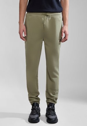 Mężczyzna ubrany w oliwkowozielone spodnie dresowe ze ściągaczem w pasie oraz czarno-szare trampki, stojący na tle jednolitego szarego tła.