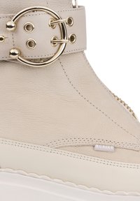 Bottine en cuir beige à forme arrondie, ornée d'une boucle circulaire en or et d'une fermeture éclair sur le côté. Semelle blanche avec bord ondulé et étiquette de marque.
