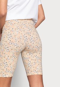 Nainen, joka pitää ylös nostettuja beigen värisiä kukkakuosisia pyöräilyshortseja ja valkoista yläosaa, kuvattuna takaa yksinkertaista vaaleaa taustaa vasten.