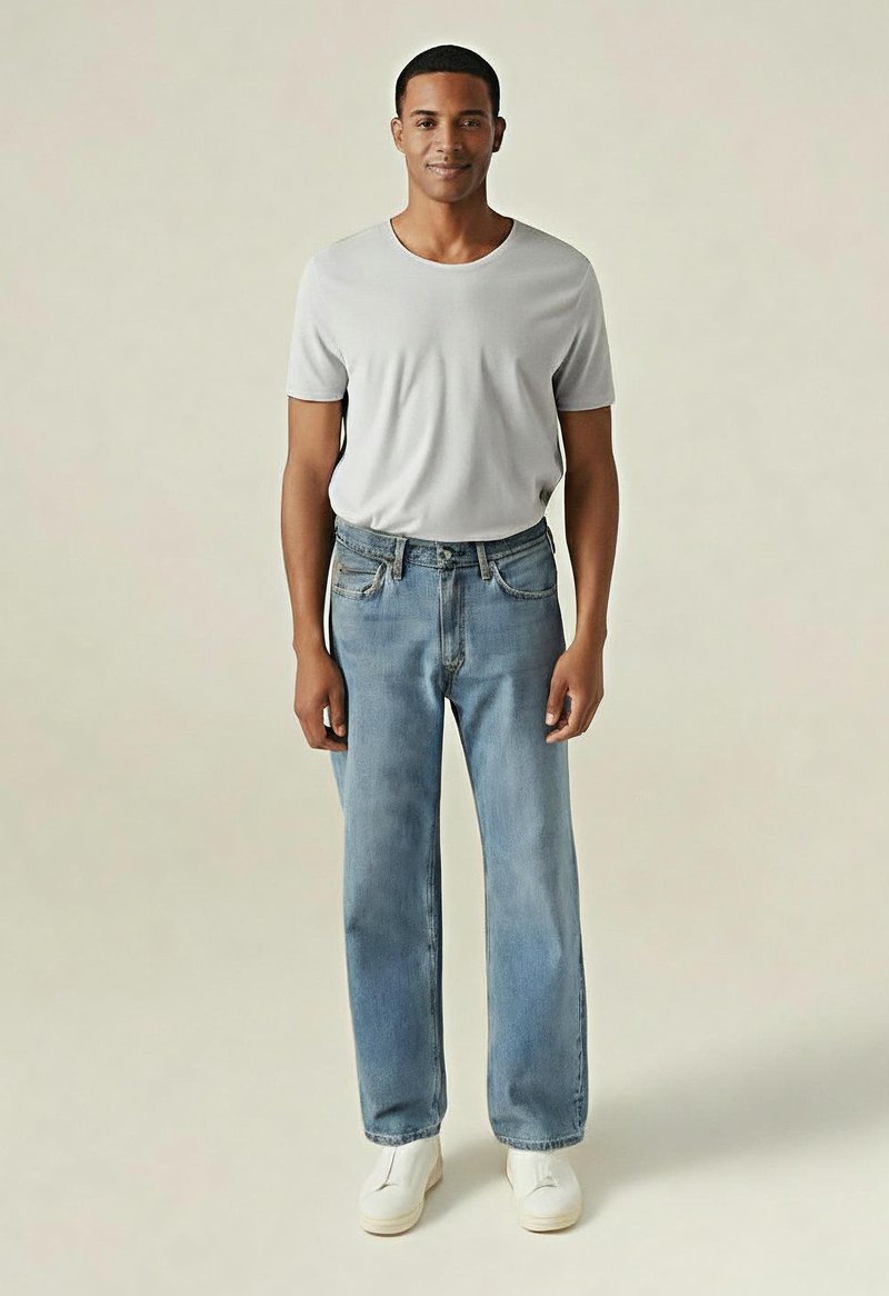 Hombre de pie de frente con una camiseta gris clara lisa, vaqueros azul claro y zapatillas blancas sin cordones, contra un fondo beige.