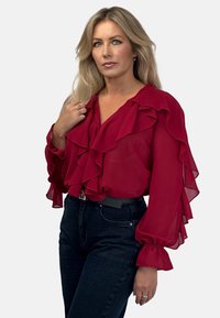 Czerwona bluzka z szyfonu z falbaniastymi detalami, dekoltem w kształcie litery V i przezroczystymi długimi rękawami. W zestawie z dżinsami z wysokim stanem w ciemnym odcieniu i czarnym paskiem.