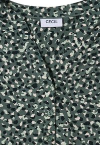 Grüne Bluse mit Knopfleiste, die ein Leopardenmuster in verschiedenen Schattierungen zeigt, mit V-Ausschnitt und strukturierter Stoffqualität. Das Etikett trägt die Aufschrift "CEcil".