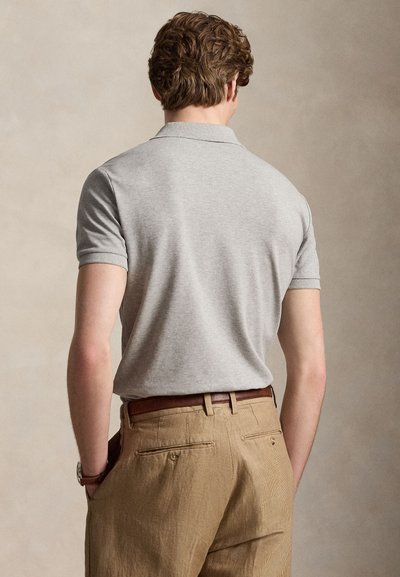 Vyras su garbanotais plaukais, dėvintis šviesiai pilką trumpomis rankovėmis polo marškinėlį, įkištą į smėlio spalvos korduro klostėtus kelnes su rudu diržu, rankos kišenėse.