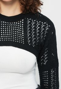 Γυναίκα που φοράει μαύρο πλεκτό μπολερό πάνω από ένα λευκό στενό τοπ, δείχνοντας το υφασμένο μοτίβο και τα κατσαρά μαλλιά σε ουδέτερο φόντο.