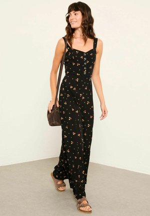 Frau geht drinnen und trägt einen schwarzen Jumpsuit mit Blumenmuster, braune Sandalen und eine braune Umhängetasche.