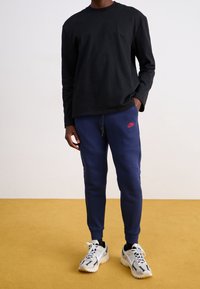 Person med svart långärmad tröja, marinblå Nike träningsbyxor med röd logotyp och vita sportskor på en gul golv mot en vit vägg.