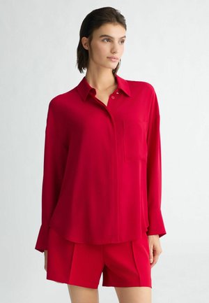 Completo composto da blusa rossa e pantaloni corti in tessuto leggero e morbido. La blusa presenta un colletto, una chiusura frontale con bottoni e una tasca sul petto.