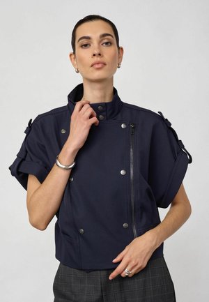Femme portant une veste bleu marine à manches courtes avec fermeture éclair et boutons-pression, associée à un pantalon à carreaux gris, posant devant un fond uni.
