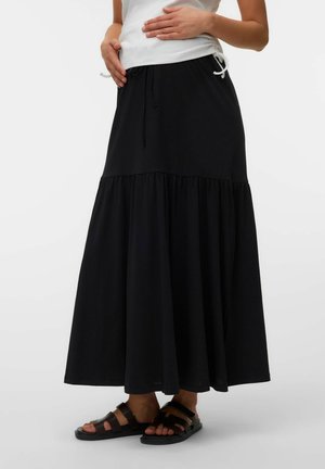 Kvinde i en lang sort lagdelt nederdel, hvid ærmeløs top og sorte sandaler, stående med hænderne på hoften.