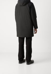 Svart, lång, puffig jacka med huva, med en slät textur. Kombinerad med svarta byxor och robusta svarta skor. Bakifrån.