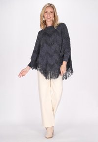 Poncho grigio scuro con motivo a zigzag e orlo in frange, abbinato a pantaloni larghi crema e stivaletti beige chiaro.