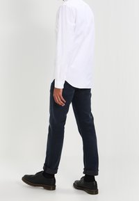 Camicia bianca a maniche lunghe, jeans in denim scuro, scarpe nere, pantaloni con risvolti, cuciture visibili sulle maniche, texture del tessuto liscia.
