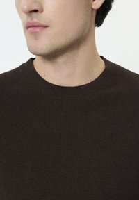 Pull marron côtelé avec un col rond. Vue en gros plan mettant en valeur la texture et le motif du tissu, montrant des détails subtils.