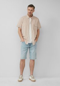 Korte mouwen geruite blouse in oranje en wit, lichtblauwe denimshorts en crèmekleurige sneakers met tan zolen. Model staat met handen in de zakken.