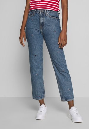 Personne portant un jean bleu taille haute avec de petits logos brodés, un haut rayé rouge et blanc, et des baskets blanches, debout devant un fond gris.
