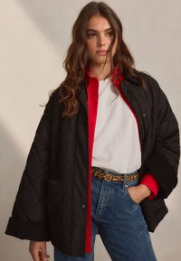 Black quilted jacket with a red interior, featuring a relaxed fit, snap buttons, and side pockets, worn over a white shirt and blue jeans.