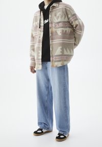 Camicia a quadri con motivi geometrici rosa, beige e grigi sopra una felpa nera, abbinata a jeans larghi di un blu chiaro e scarpe con dettagli neri.