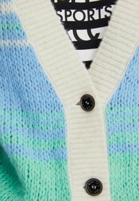 Knitted cardigan featuring a blend of blue and green stripes, with a cream-colored placket and black buttons. Textured fabric detail visible.