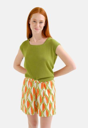 Joven mujer con cabello rojo que lleva una camiseta verde de manga corta y pantalones cortos con estampado naranja, verde y blanco, con las manos apoyadas en las caderas.
