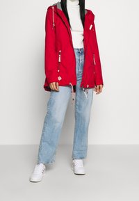 Červená voděodolná bunda se šedou kapucí, stahovacími šňůrkami a kapsami na zip. Nositá přes bílý rolák a světlé modré džíny. Bílé tenisky.