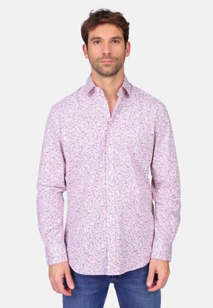 Homme debout face à l'avant, portant une chemise à manches longues rose à motifs floraux et un jean bleu, sur un fond uni.