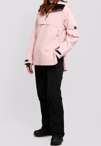 Giacca impermeabile rosa chiaro con cappuccio nero, tasca frontale e dettagli con cerniera, abbinata a pantaloni neri e calzature nere.