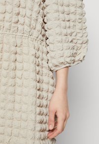 Μπεζ φόρεμα με υφή που διαθέτει ανώμαλη επιφάνεια, φουσκωτά μανίκια και σφικτή μέση, αναδεικνύοντας ένα ξεχωριστό σχέδιο και μαλακό υλικό.