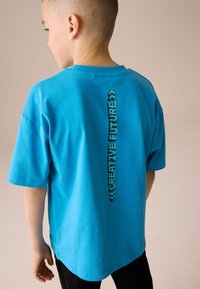 Turquoise katoenen T-shirt met korte mouwen en een rechte zoom. Op de achterkant staat in contrasterende donkerblauwe letters "CREATIVE FUTURE".
