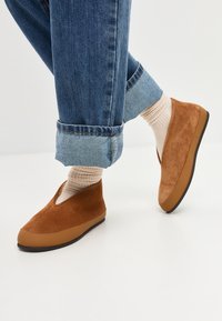 Bruine suède instapschoenen met een ronde neus en een rubberen zool. Gedingd bij opgerolde spijkerbroek en lichtgekleurde geribde sokken.