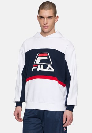 Homme portant un hoodie FILA blanc, marine et rouge et un short en maille navy, se tenant devant un fond gris clair uni.