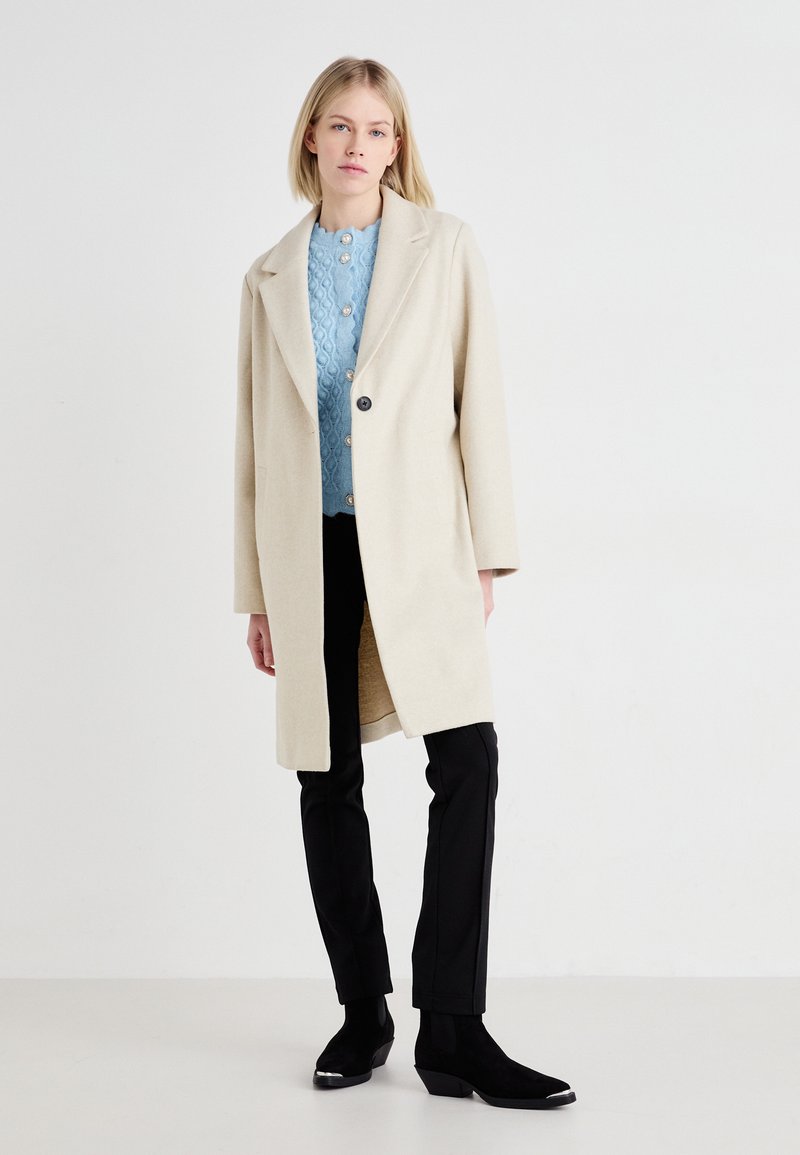 Beige Wollmantel mit breitem Revers, offenem Front und weicher Textur, kombiniert mit einem hellblauen strukturierten Pullover und schwarzen, eng anliegenden Hosen.