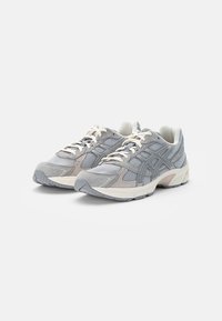 Zapatillas deportivas grises con parte superior de malla y ante, cordones blancos y suela acolchada. Presenta una marca destacada y detalles texturizados.