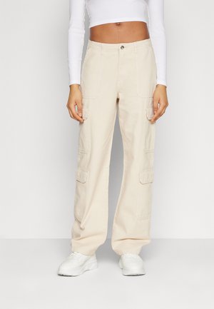 Pantalons cargo beige à coupe ample, dotés de plusieurs poches à patch, d'une fermeture à bouton et d'une coupe droite. Associés à des baskets blanches.
