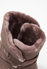 Botas de ante marrón con un forro suave y esponjoso de piel de color marrón claro. El puño está enrollado, revelando la textura interior. Presenta un detalle de botón en el lateral.