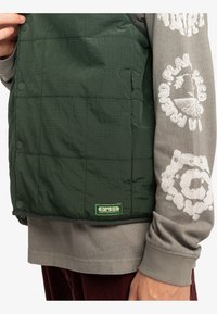 Groene gewatteerde vest met drukknoopsluitingen en een klein voorvak, gecombineerd met een grijs shirt met lange mouwen met bedrukt detail.